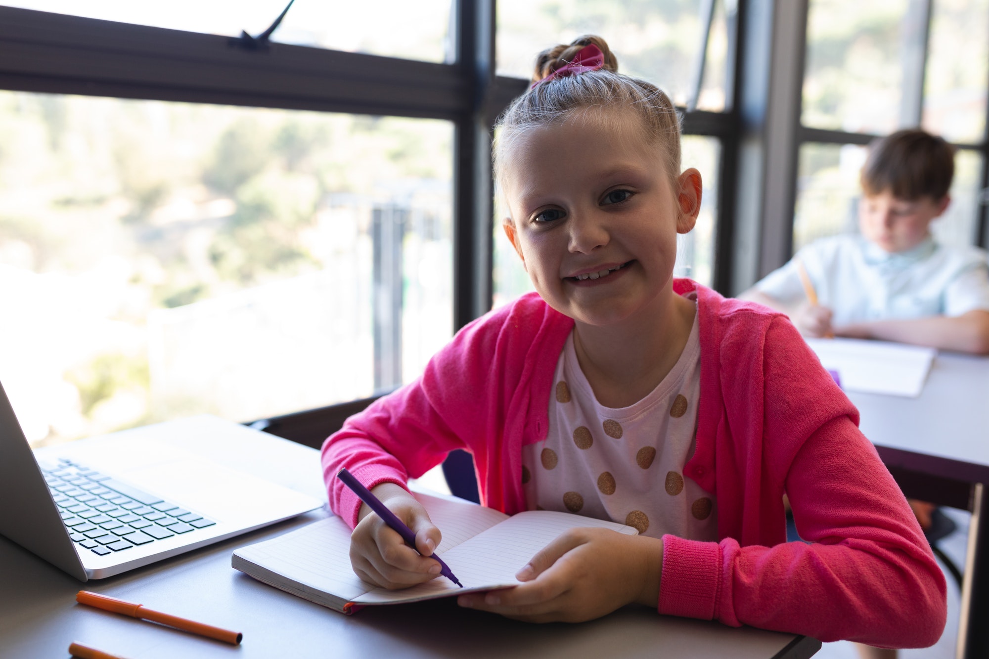 Schoolgirl writing on notebook and looking at camera at desk in classroom of elementary school
